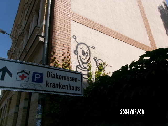 Graffiti mit fröhlichem Alien auf beiger Wand; daneben Krankenhausschild. Grüne Pflanzen im Vordergrund.