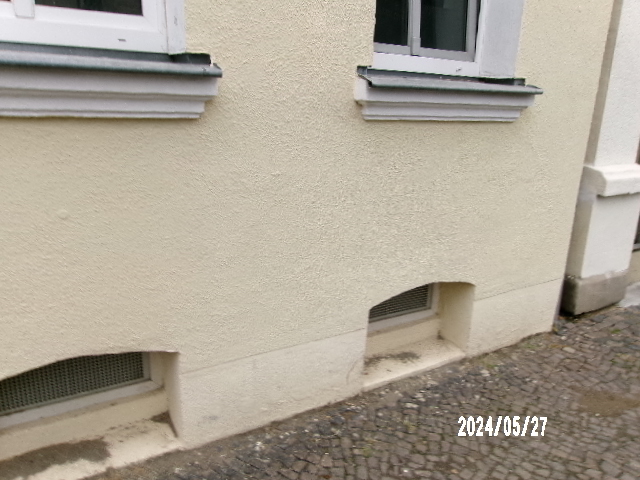 Hauswand mit zwei Fenstern und Kellerlüftungsschlitzen, helle Fassade, gepflasterter Gehweg.