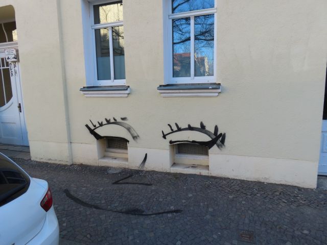 Graffiti von Augen unter Fenstern auf beiger Hauswand, humorvoll mit Wimpern, Schwarzweiß.